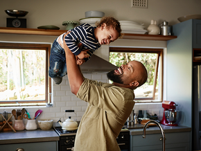 Photograph: smiling man lifts laughing toddler above him in sunlit kitchen