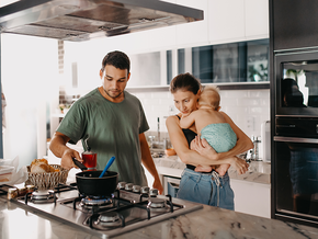 Photograph of smiling parents in modern kitchen; father cooks while mother holds toddler.