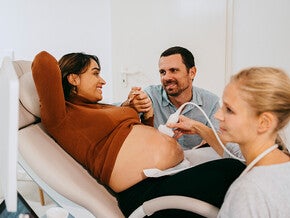 Photo: pregnant woman on exam chair, partner holding her hand, sonographer at console.