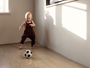 Photograph: barefoot toddler in burgundy romper kicking soccer ball on wooden floor