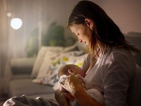 Mãe a embalar bebé ao colo num quarto pouco iluminado, possivelmente noite. Bebé inquieto, mãos pequenas crispadas