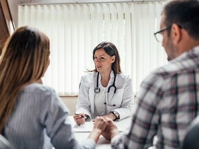 Pediatra de bata branca a explicar algo aos pais numa consulta, gesto de mão calmo, pais atentos de costas/perfil