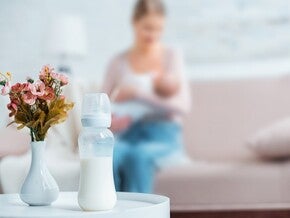 Mãe a amamentar bebé num sofá, com biberão preparado pousado na mesa lateral. Coexistência peito + fórmula sem julgamento