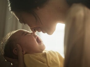 Mãe com bebé ao colo junto a uma janela, a olhar para ele com calma. Luz natural suave, tom científico acessível. Sem biberão nem produto visível, sem logótipos.