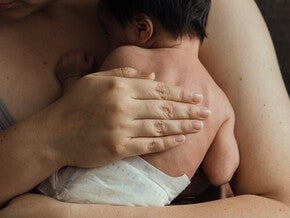 Mão adulta a segurar a mão minúscula de um bebé prematuro. Cuidado pele com pele (método canguru). Tom íntimo e sensível, luz quente e ténue. Sem logótipos.