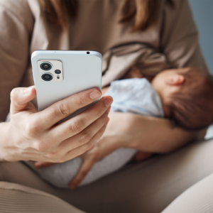 Photograph: adult holding smartphone while cradling a sleeping newborn wrapped in light blanket.