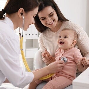Mãe a conversar com a pediatra em consulta, ambas inclinadas para o bebé. Tom de decisão informada e acompanhada, iluminação natural. Sem produto visível, sem logótipos.