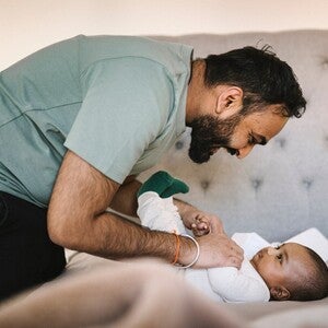 Pai sorrindo brinca com bebê deitado em cama cinza.