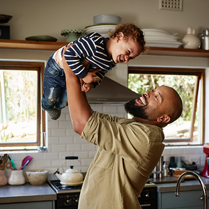 Photograph: smiling man lifts laughing toddler above him in sunlit kitchen