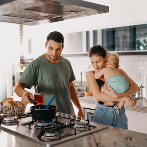 Photograph of smiling parents in modern kitchen; father cooks while mother holds toddler.