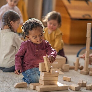 Photograph: young child focused building wooden blocks on carpet while others play nearby