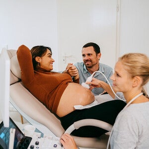 Photo: pregnant woman on exam chair, partner holding her hand, sonographer at console.