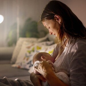Mãe a embalar bebé ao colo num quarto pouco iluminado, possivelmente noite. Bebé inquieto, mãos pequenas crispadas