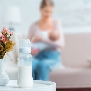 Mãe a amamentar bebé num sofá, com biberão preparado pousado na mesa lateral. Coexistência peito + fórmula sem julgamento