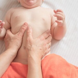 Mão de adulto a fazer massagem suave em barriga de bebé deitado sobre uma manta de algodão