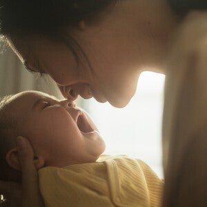 Mãe com bebé ao colo junto a uma janela, a olhar para ele com calma. Luz natural suave, tom científico acessível. Sem biberão nem produto visível, sem logótipos.