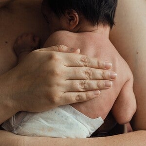 Mão adulta a segurar a mão minúscula de um bebé prematuro. Cuidado pele com pele (método canguru). Tom íntimo e sensível, luz quente e ténue. Sem logótipos.