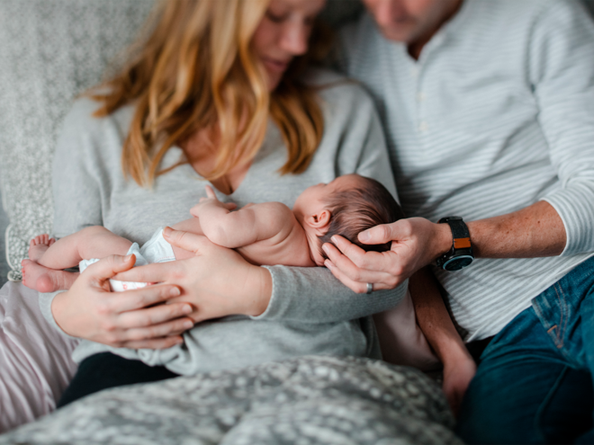 Photograph of parents gently cradling a newborn on a gray couch