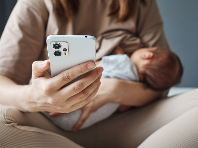 Photo: woman cradling sleeping baby while checking smartphone