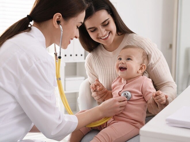 Mãe a conversar com a pediatra em consulta, ambas inclinadas para o bebé. Tom de decisão informada e acompanhada, iluminação natural. Sem produto visível, sem logótipos.
