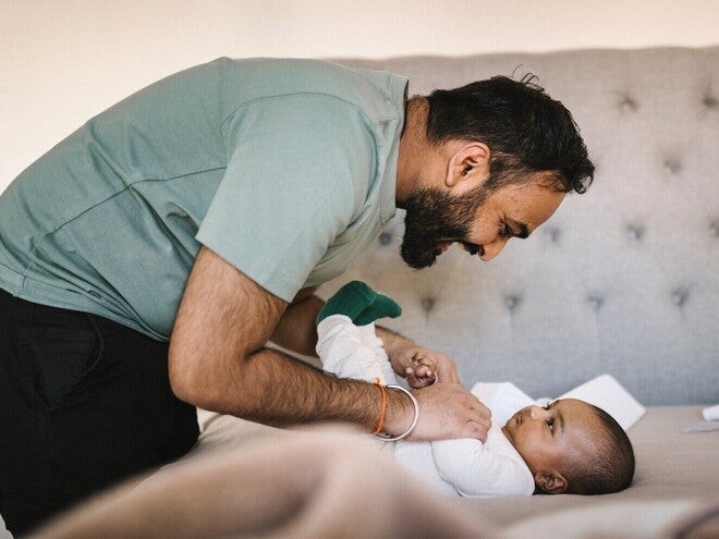 Pai sorrindo brinca com bebê deitado em cama cinza.