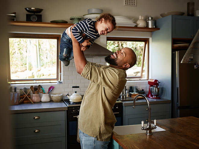 Photograph: smiling man lifts laughing toddler above him in sunlit kitchen