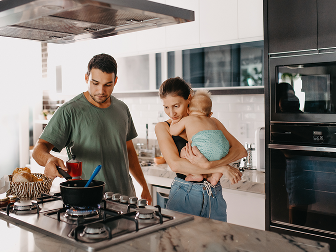 Photograph of smiling parents in modern kitchen; father cooks while mother holds toddler.