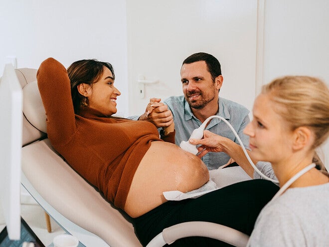 Photograph: pregnant woman on exam chair, partner holding her hand, sonographer at ultrasound