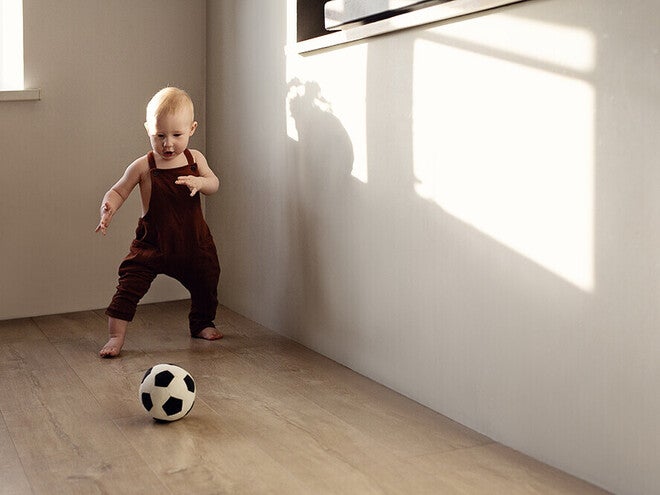 Photograph: barefoot toddler in burgundy romper kicking soccer ball on wooden floor