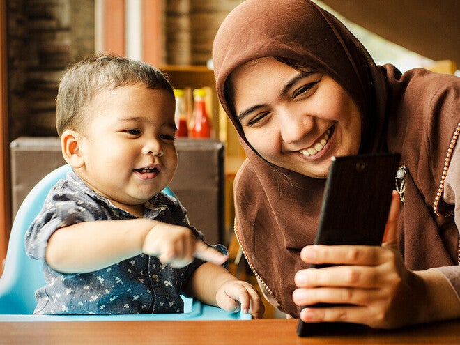 Photograph of smiling woman in hijab showing smartphone to toddler in a high chair