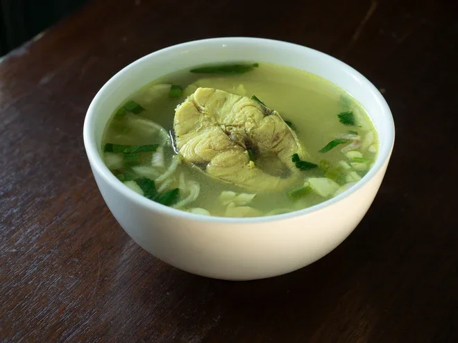 Photograph: white bowl of clear fish soup with herbs on wooden table
