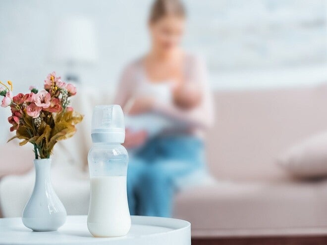 Mãe a amamentar bebé num sofá, com biberão preparado pousado na mesa lateral. Coexistência peito + fórmula sem julgamento