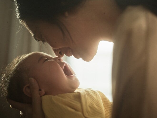 Mãe com bebé ao colo junto a uma janela, a olhar para ele com calma. Luz natural suave, tom científico acessível. Sem biberão nem produto visível, sem logótipos.