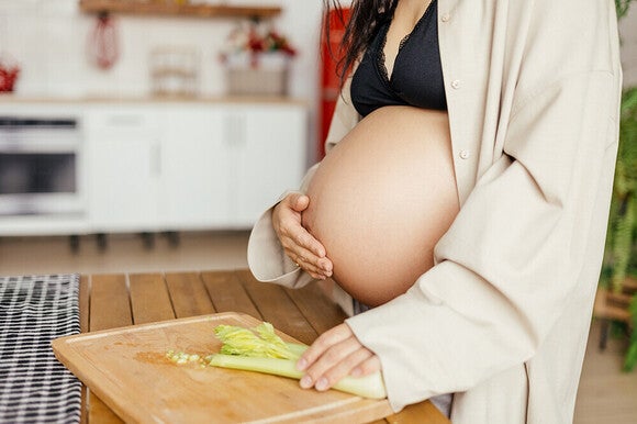 Photograph of pregnant woman in robe holding belly while cutting celery on wooden board