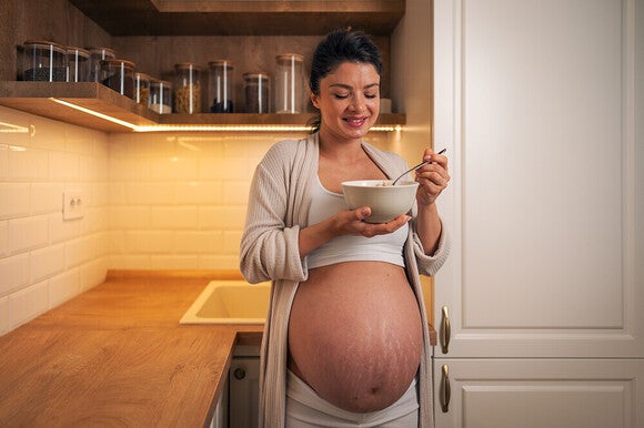 Photograph of smiling pregnant woman with exposed belly eating from a bowl in warm kitchen